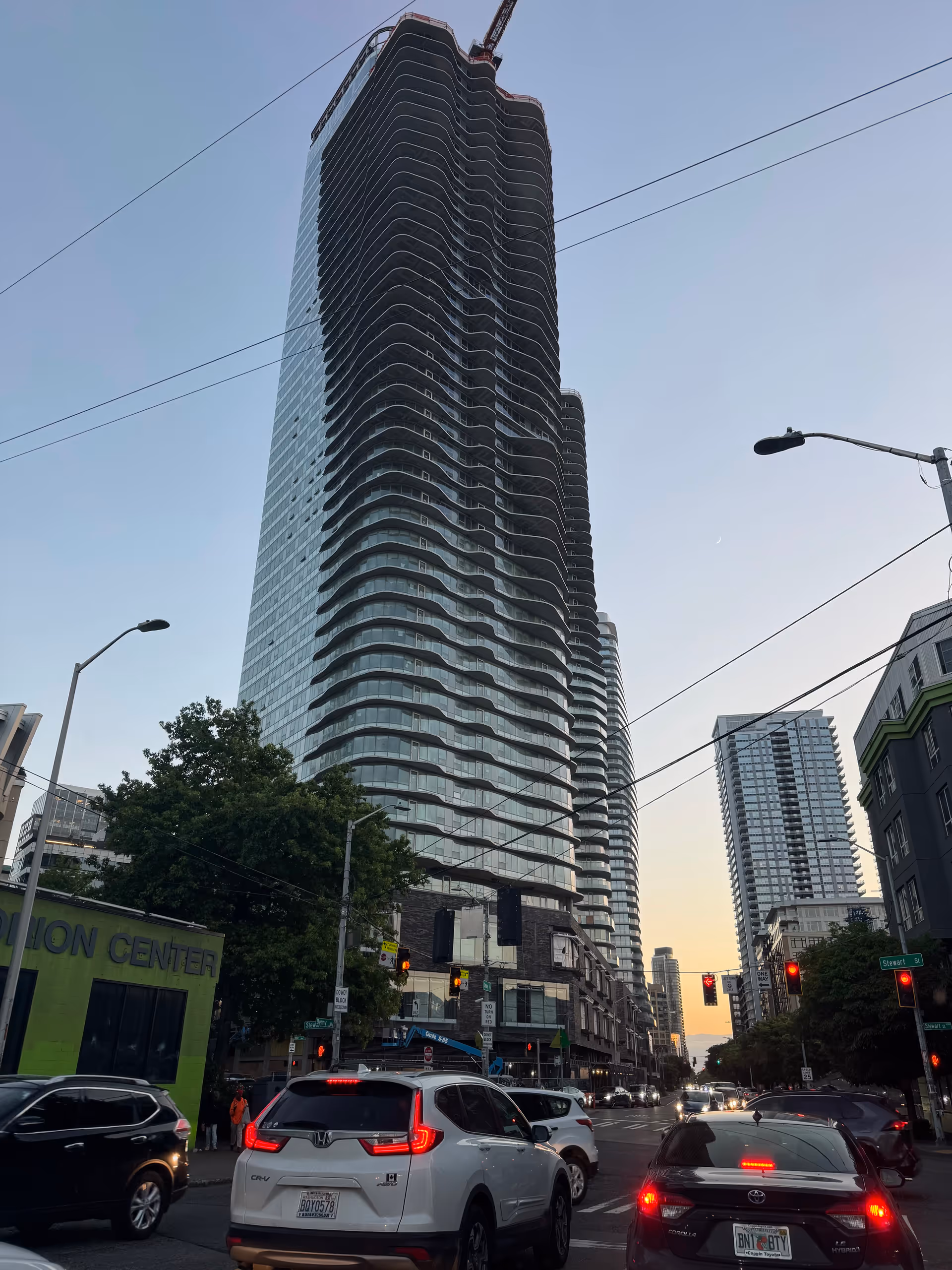 Tall modern high-rise under construction towers over a busy urban street with cars.