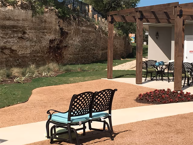 Outdoor seating area with wrought-iron benches, a table under a pergola beside a rocky wall.