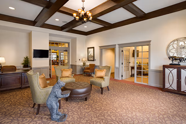 Lobby seating area in an assisted living facility with armchairs around a round ottoman, a coffered ceiling, and a small bear statue holding a bowl.