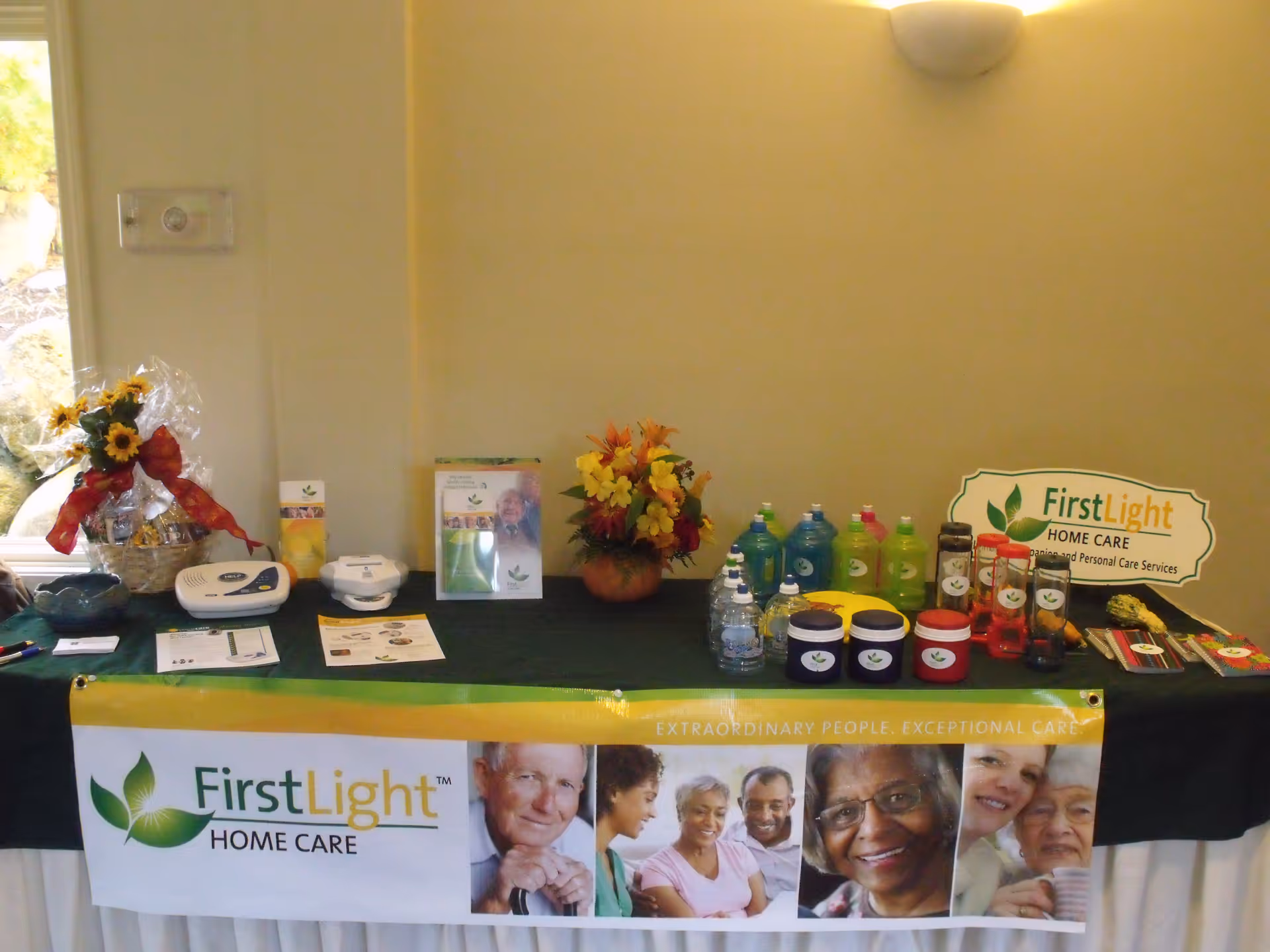 Indoor promotional display table for FirstLight Home Care with banners, flowers, and products.