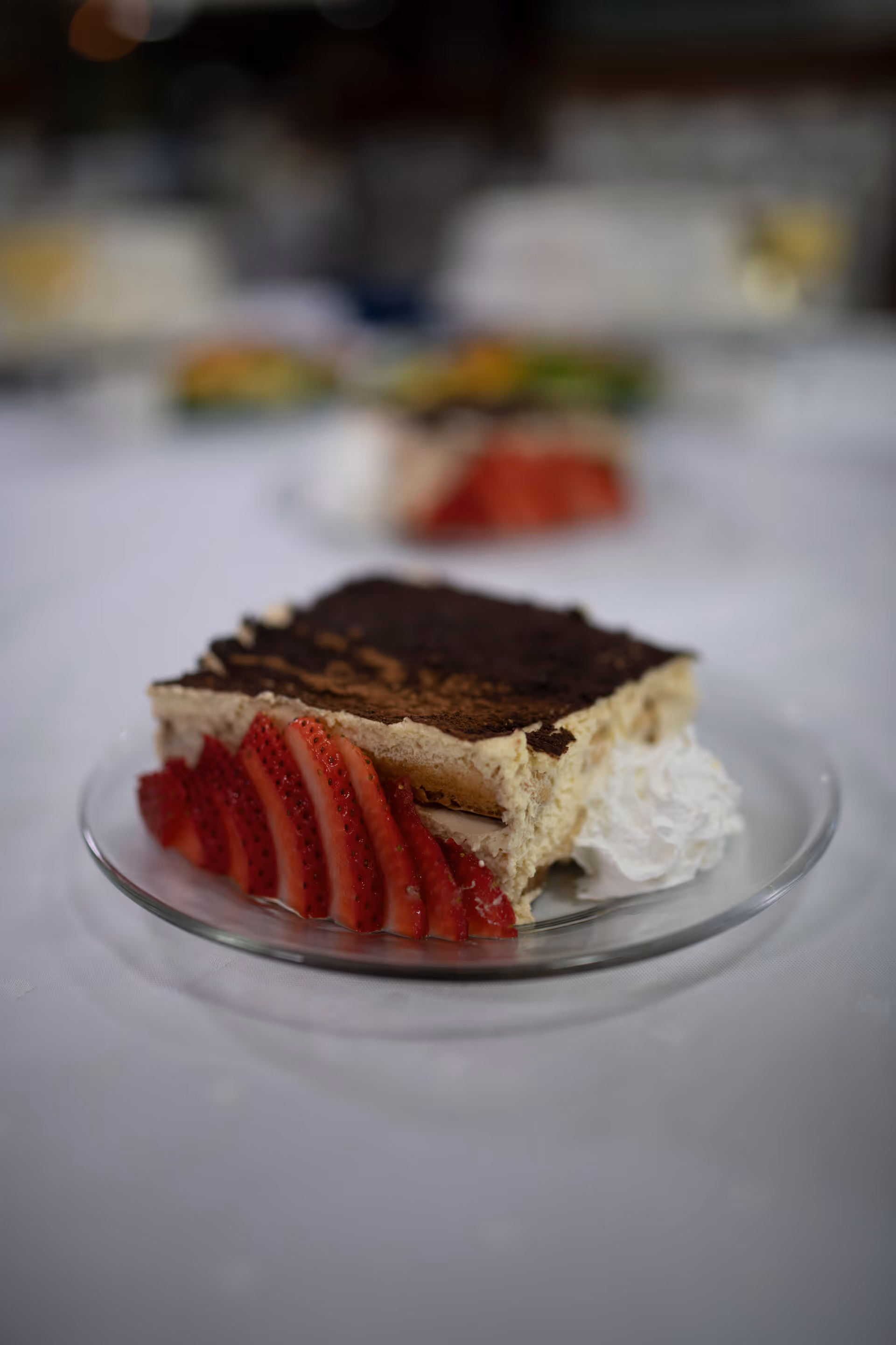 Dessert plate with cake and strawberry slices on white tablecloth; dining area blurred.