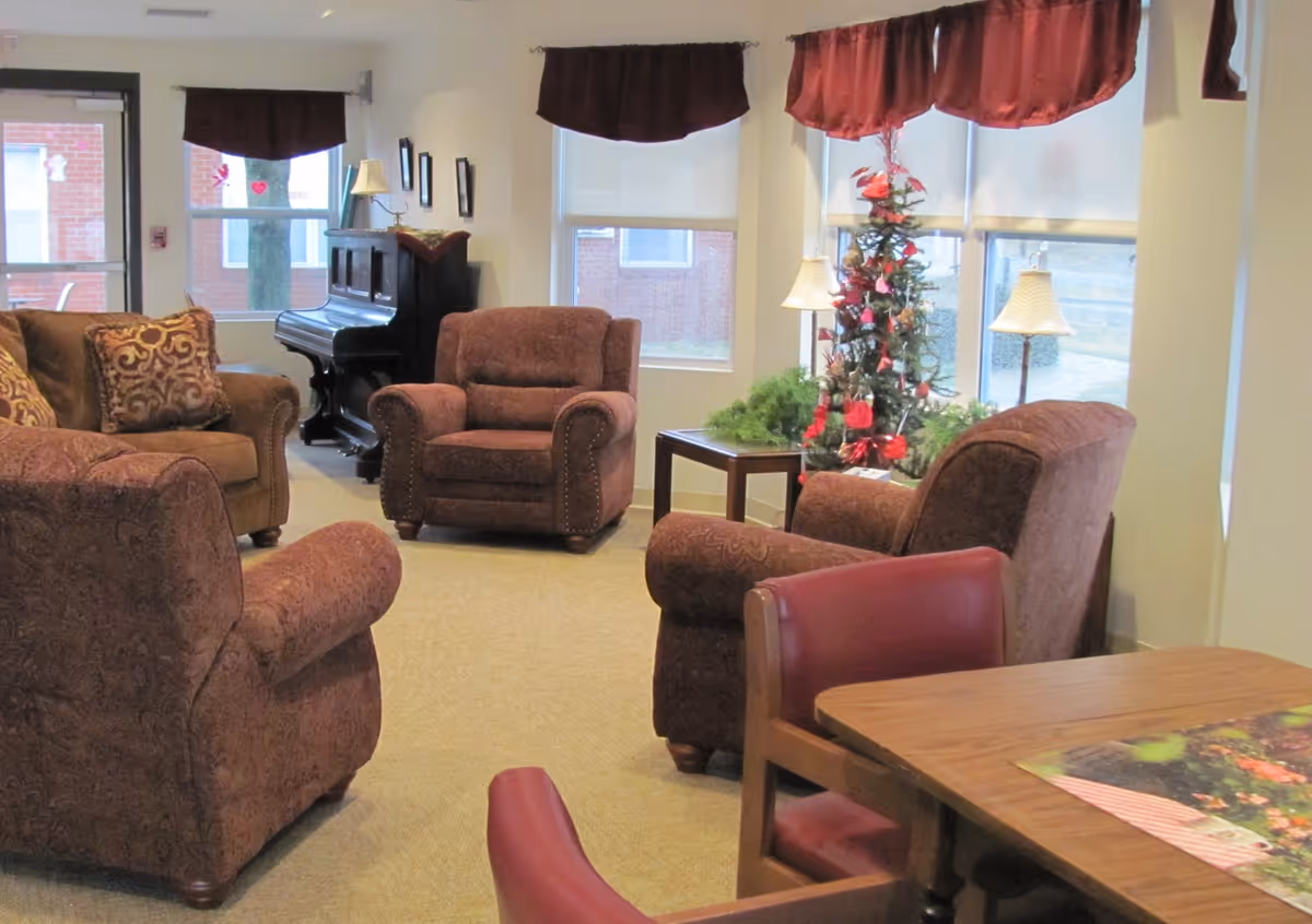 A cozy living room area with several upholstered armchairs and a sofa arranged around a small table. There is a decorated Christmas tree near the windows, a piano against the far wall, and a wooden table with red chairs in the foreground. The room has large windows with maroon valances and beige carpeted flooring.