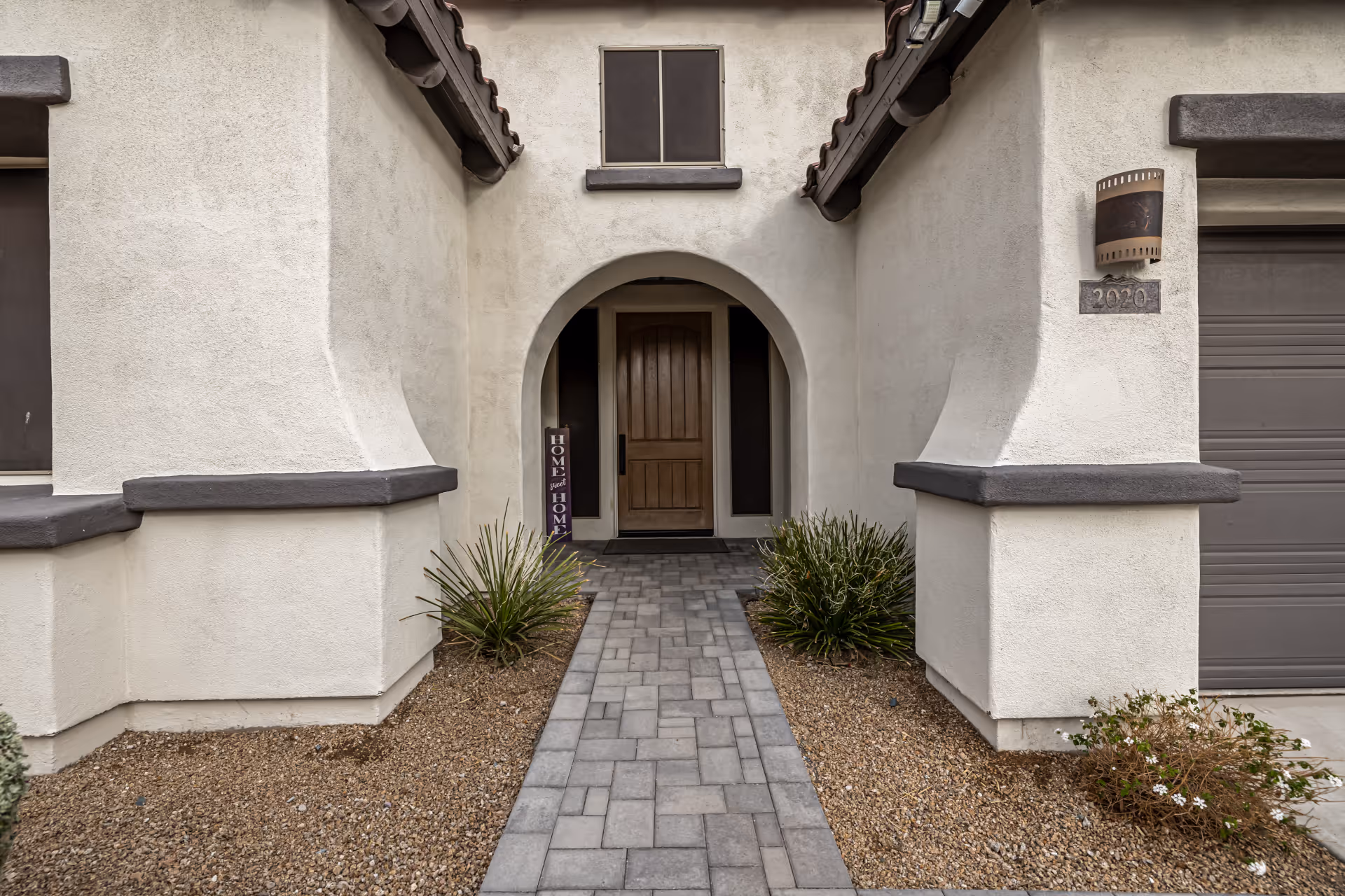 Front entrance of a stucco house with an arched doorway, paved walkway, and a garage to the right.