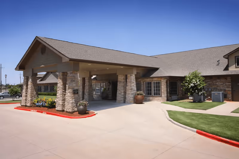 Exterior view of Legacy Ranch facility entrance with a covered drop-off area supported by stone pillars, surrounded by landscaped greenery and a clear blue sky.