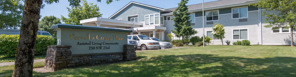 Front view of Corvallis Caring Place assisted living building with a stone sign, lawn, entrance canopy and parked vehicles.