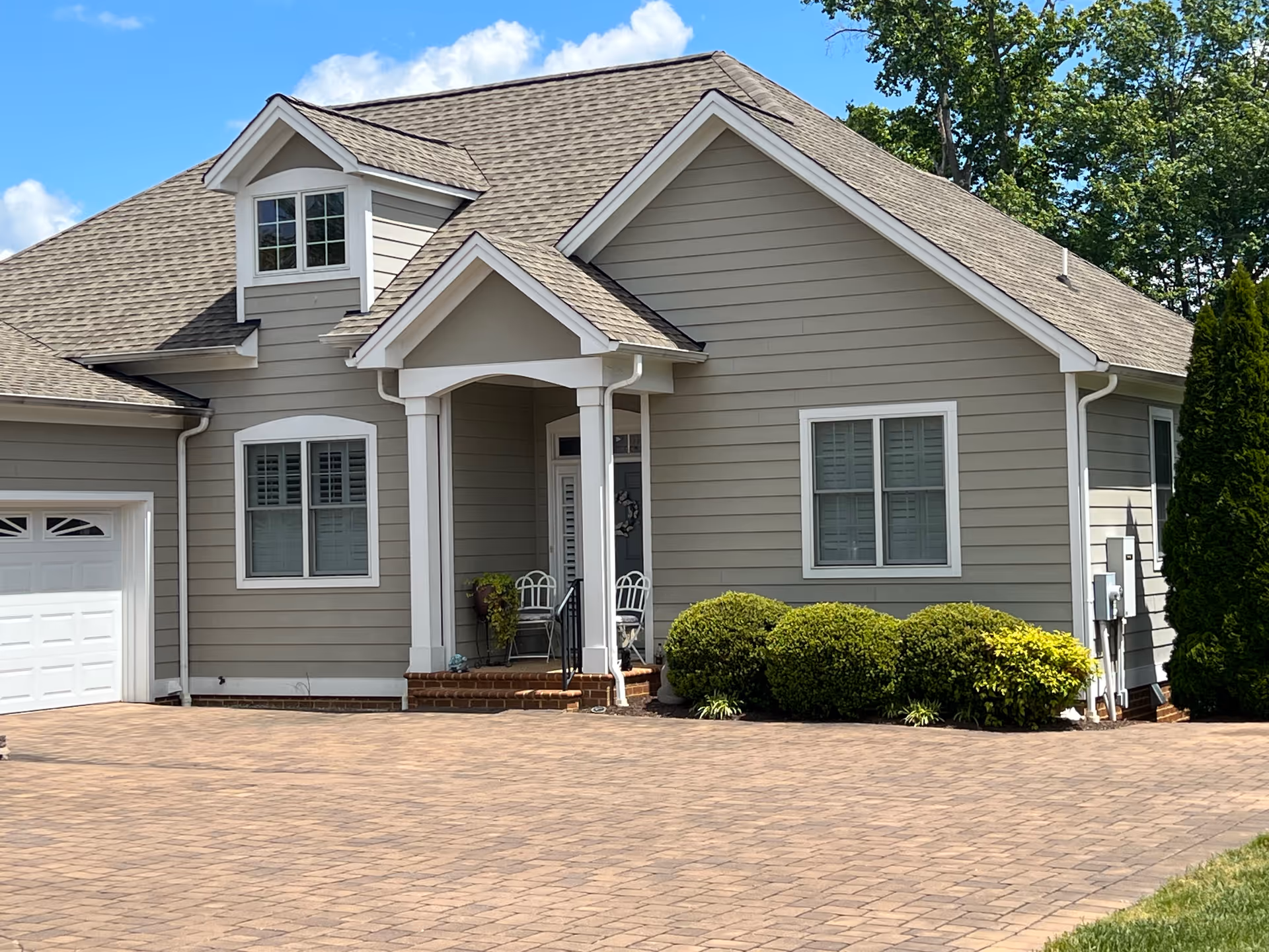 Exterior view of a single-story residential building with gray siding, white trim, and a shingled roof. The house features a small covered porch with two white chairs and some potted plants. There are two windows with white shutters and a garage door on the left side. The driveway is paved with brick-like stones, and there are neatly trimmed bushes along the front of the house. Trees and a blue sky with some clouds are visible in the background.