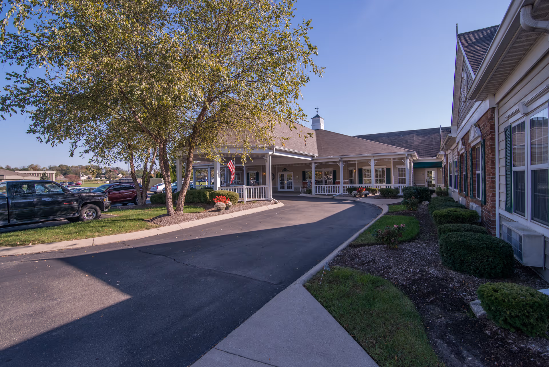 Exterior view of Morning Pointe of Franklin senior living facility showing a driveway leading to a covered entrance with white railings, surrounded by landscaped bushes and trees under a clear blue sky.