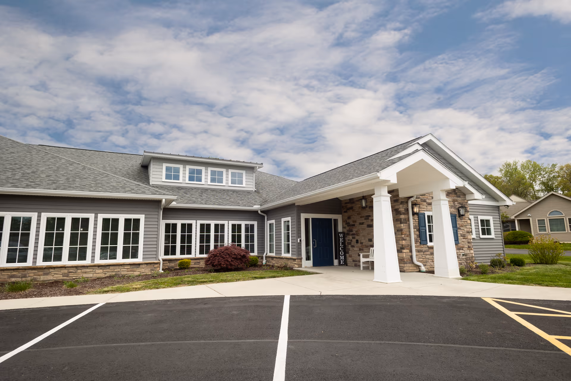Front entrance of a gray single-story senior living building with a covered portico, white columns, multiple windows, and a parking area.