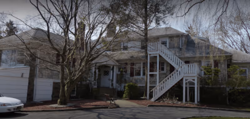 Exterior view of a two-story stone building with a white staircase on the outside leading to the upper floor. There are leafless trees in front of the building, a bench near the entrance, and a white car parked on the left side. The sky is clear and the area appears to be well-maintained.