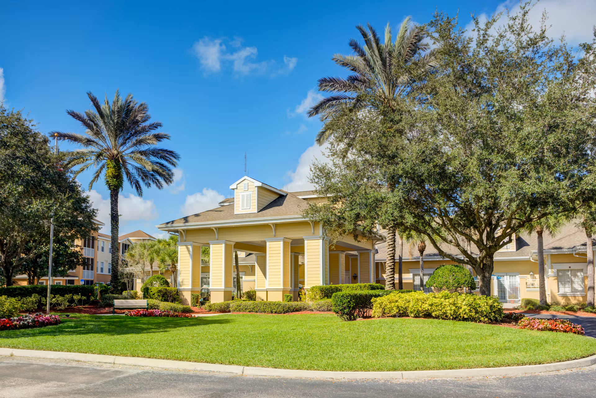 Yellow senior living facility exterior with manicured lawn, palm trees, and entrance porch.