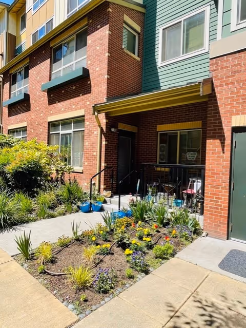 Ground-floor entrance to a brick and green-paneled building with a small landscaped garden and a patio area.