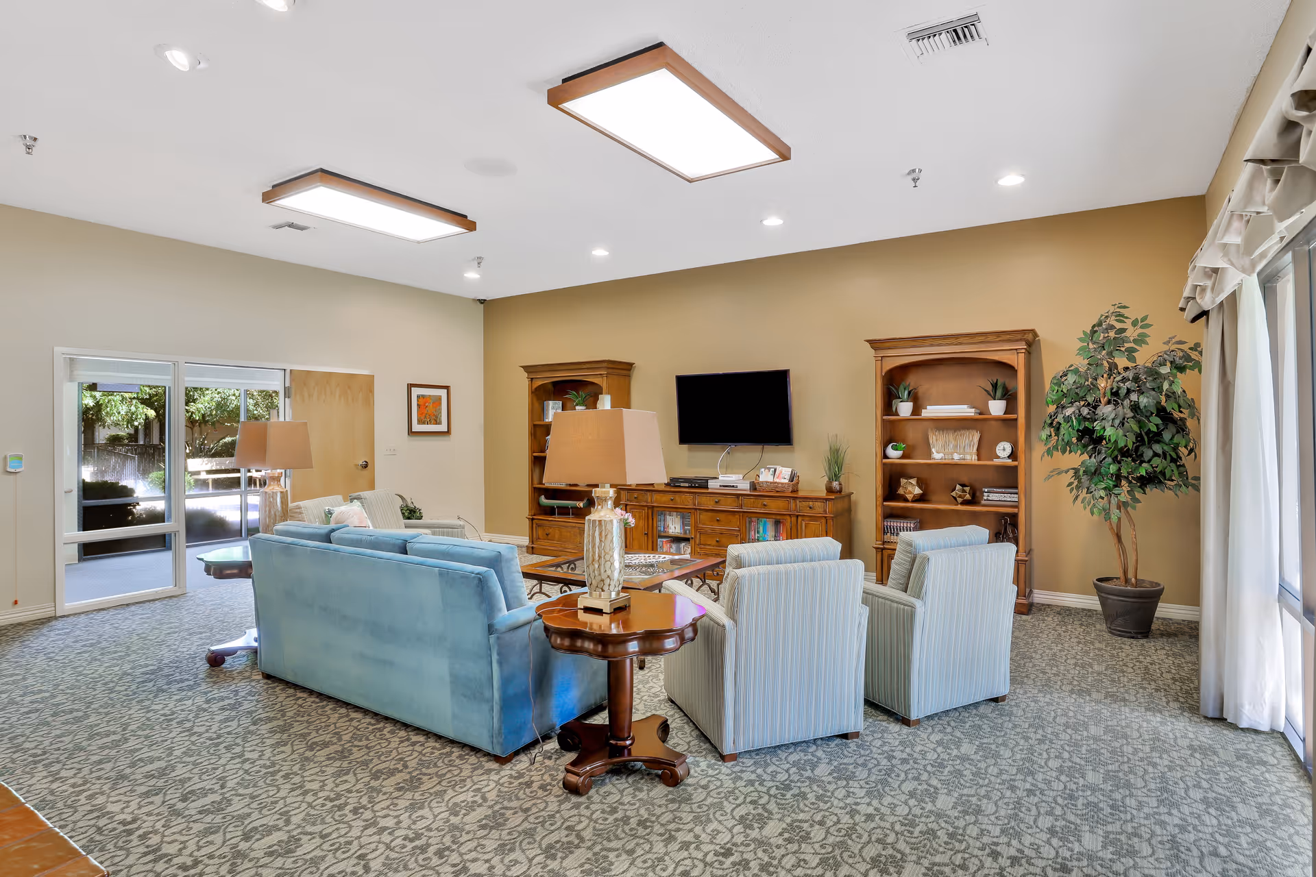 Spacious senior living lounge with blue sofa, armchairs, TV, bookcases, and plants.