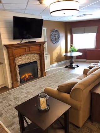 Cozy living room area with a beige sofa, a wooden coffee table with a candle lantern, a fireplace with a mounted flat-screen TV above it, a decorative wall piece, and a window with a red valance letting in natural light.