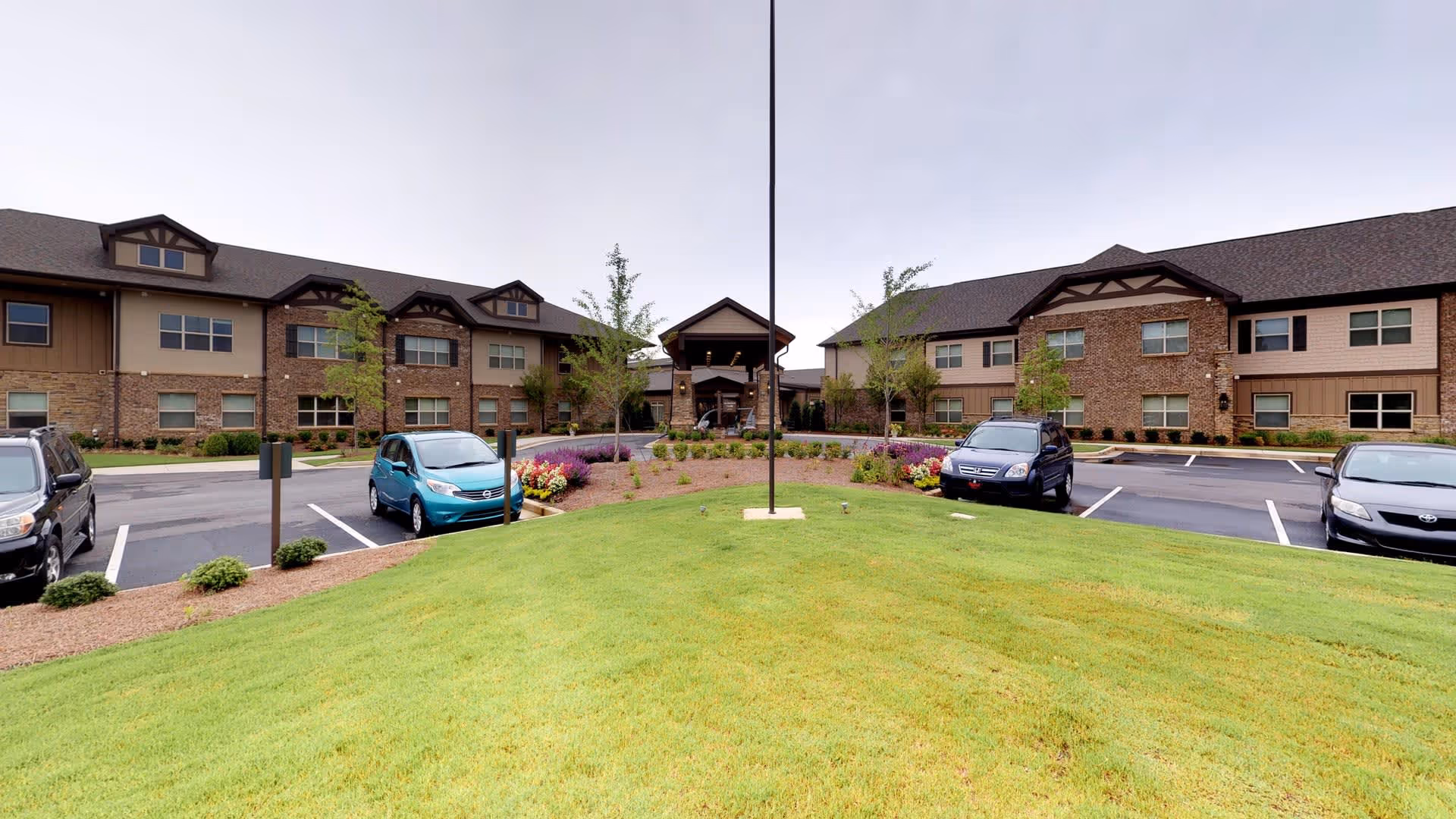 Front exterior view of Celebration Village Forsyth, showing a two-story building with brick and siding facade, a central entrance with a covered porch, a well-maintained lawn, flower beds, and a parking lot with several cars.
