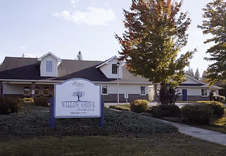 Single-story senior living facility exterior with a front lawn, walkway, trees, and a prominent "Willow Grove" sign.