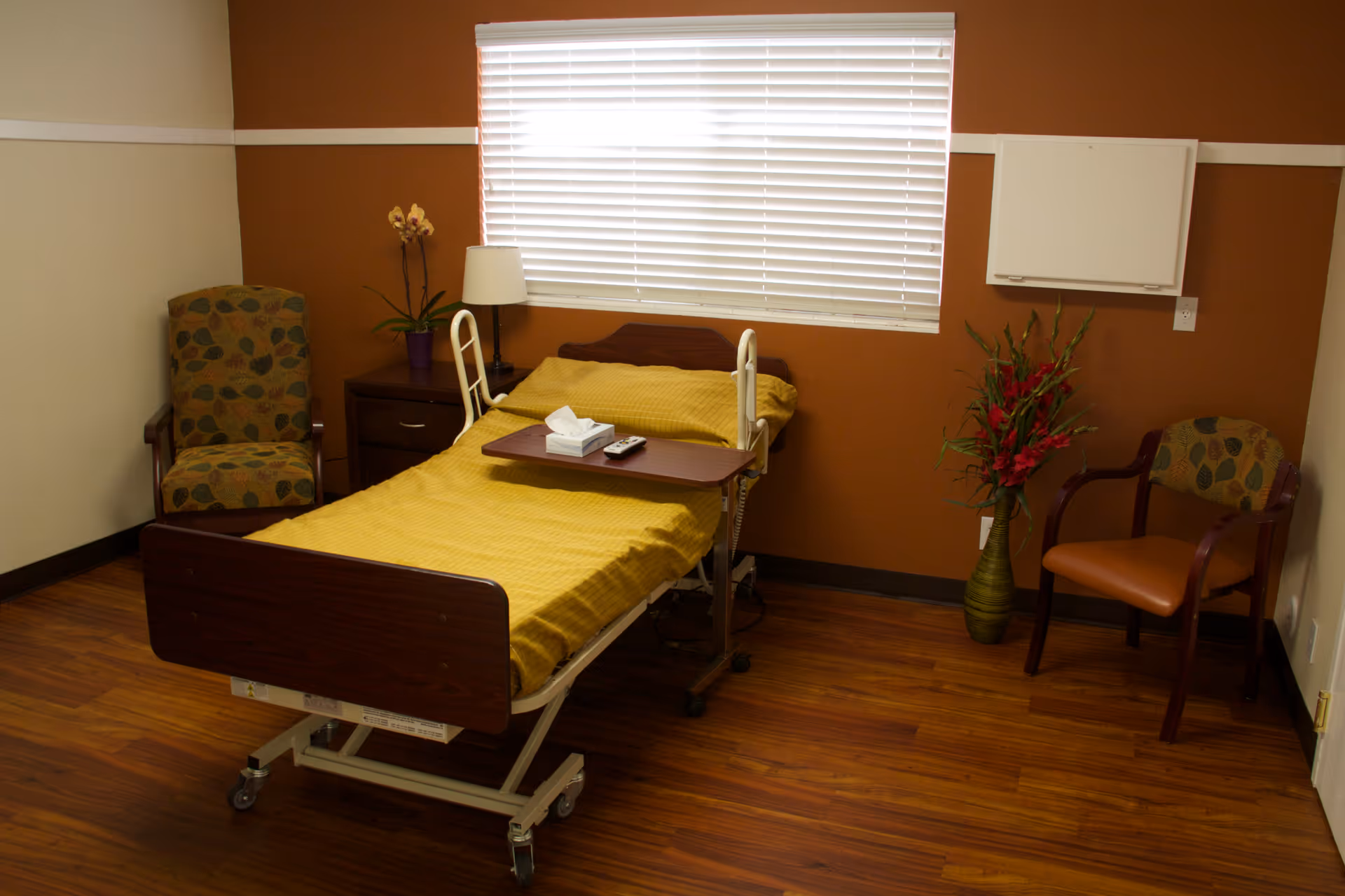 A patient room with a hospital bed, yellow bedding, chairs, and plants.