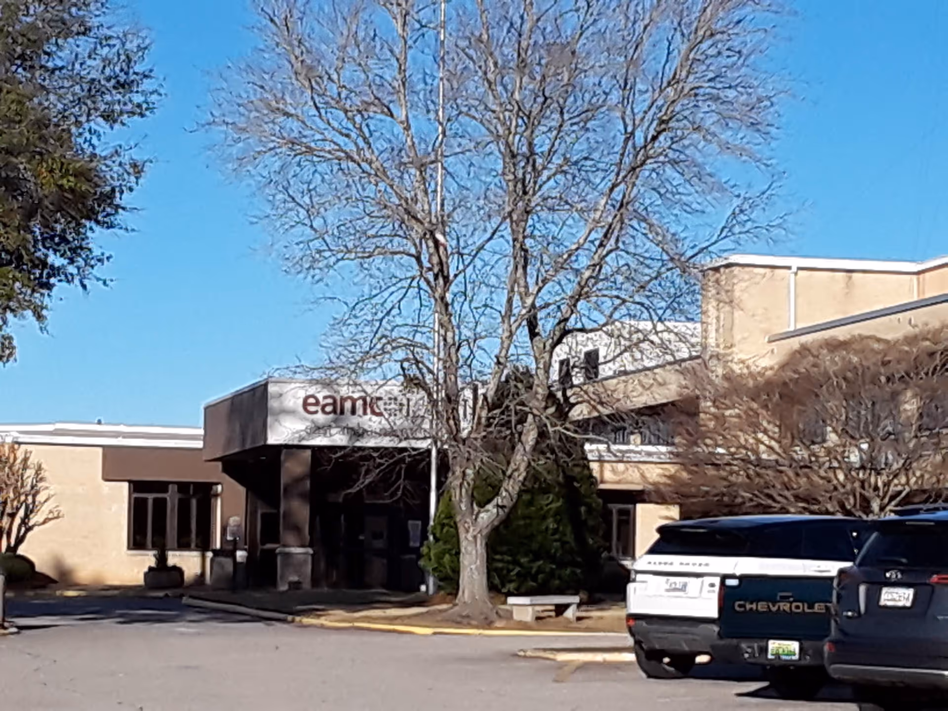 Exterior view of a beige facility entrance with parked cars and a bare tree.