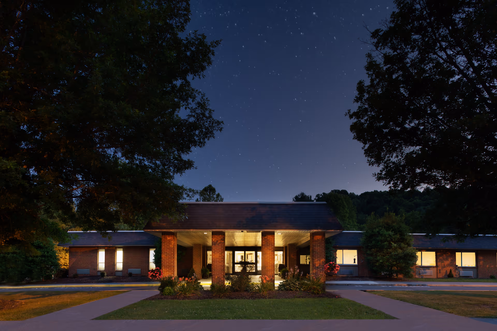 Night view of a single-story brick building with a covered entrance supported by four pillars, surrounded by trees and illuminated windows, under a starry sky.