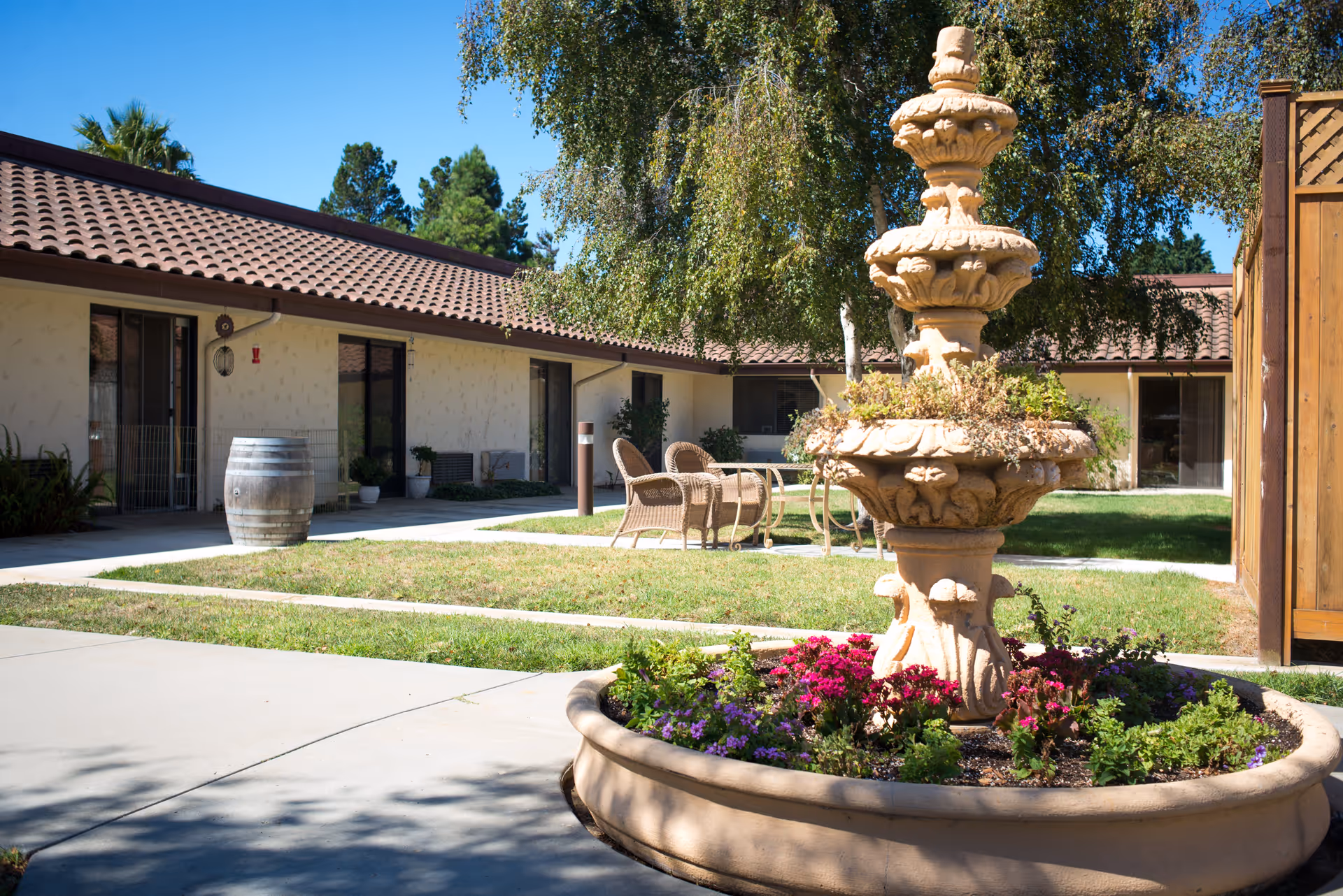 Outdoor courtyard area of a senior living facility with a decorative stone fountain surrounded by colorful flowers in the foreground, wicker chairs and a table on a patio, and a single-story building with sliding glass doors and a tiled roof in the background under a clear blue sky.