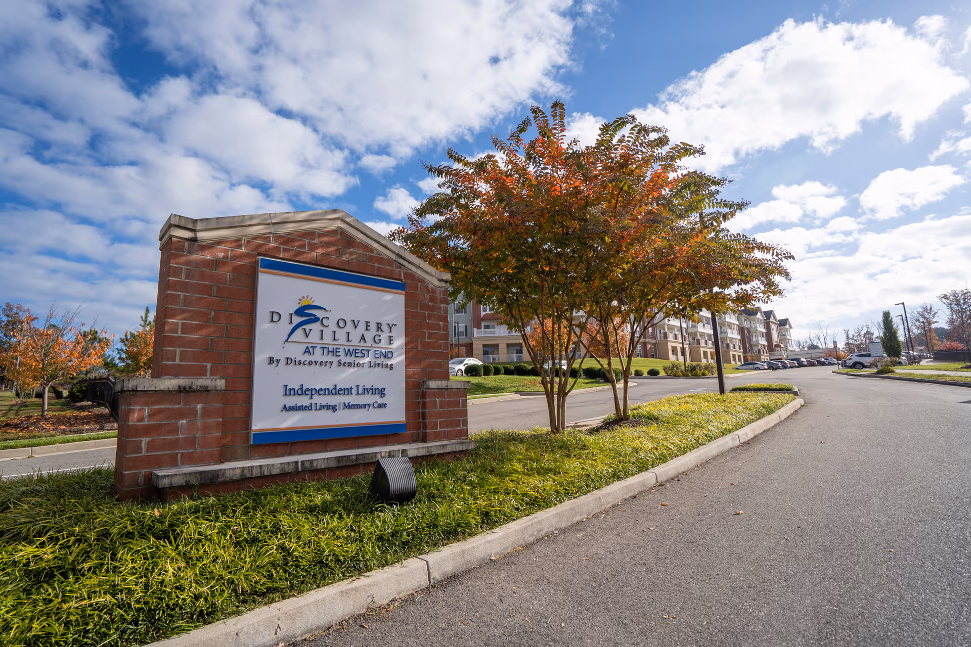 Entrance sign for Discovery Village At the West End senior living community, with a brick base and a landscaped area with grass and trees showing autumn colors. In the background, there is a paved road leading to a multi-story residential building under a partly cloudy sky.