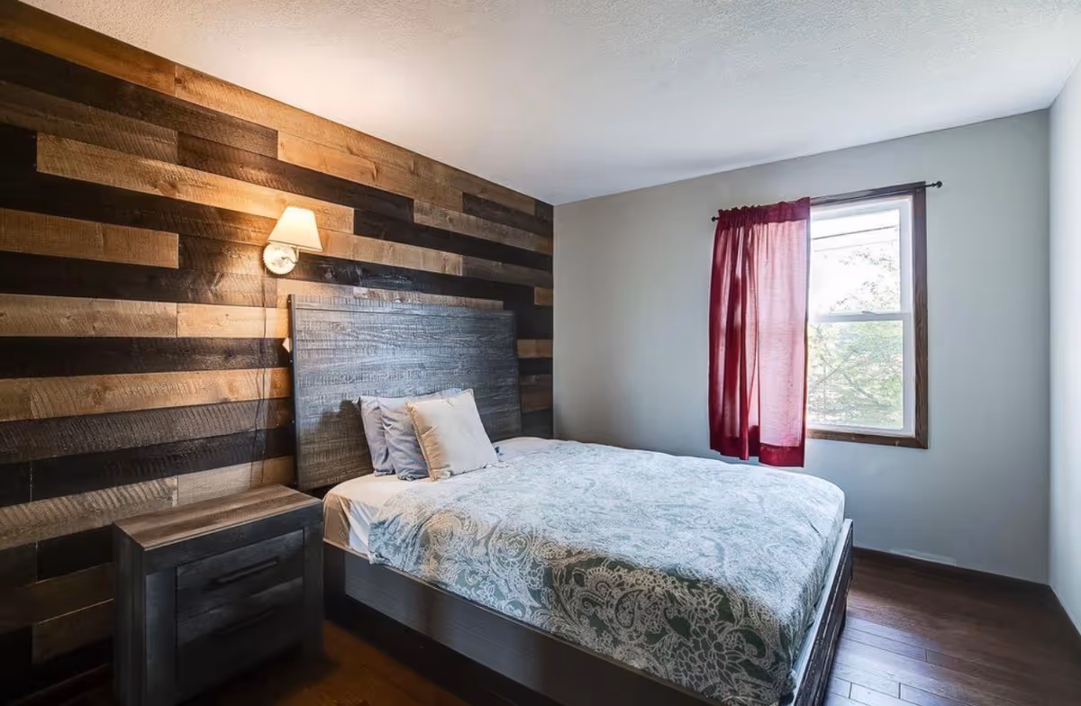 Bedroom with a striped wooden accent wall, bed, nightstand, and a window with red curtain.