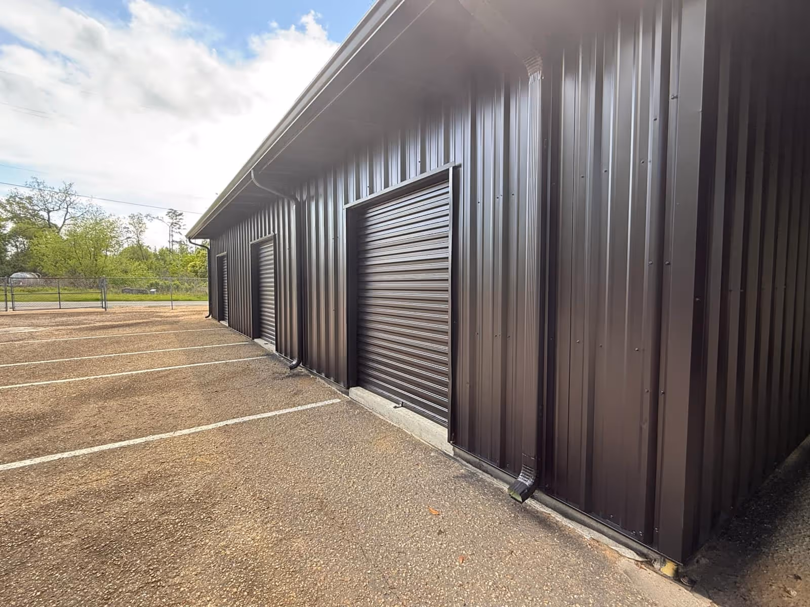 Exterior view of a dark metal building with large rolling garage doors beside a parking lot.