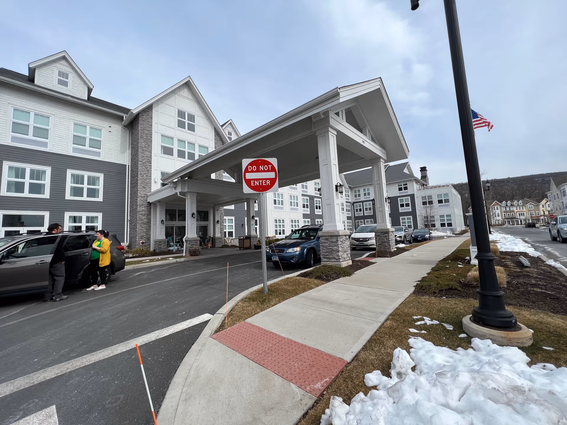 Exterior view of Skybrooke at Simsbury entrance with porte-cochere, parked cars, and pedestrians.