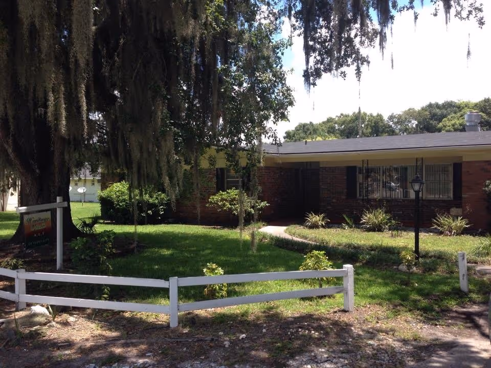 A single-story brick senior facility exterior with a manicured lawn and white fence.