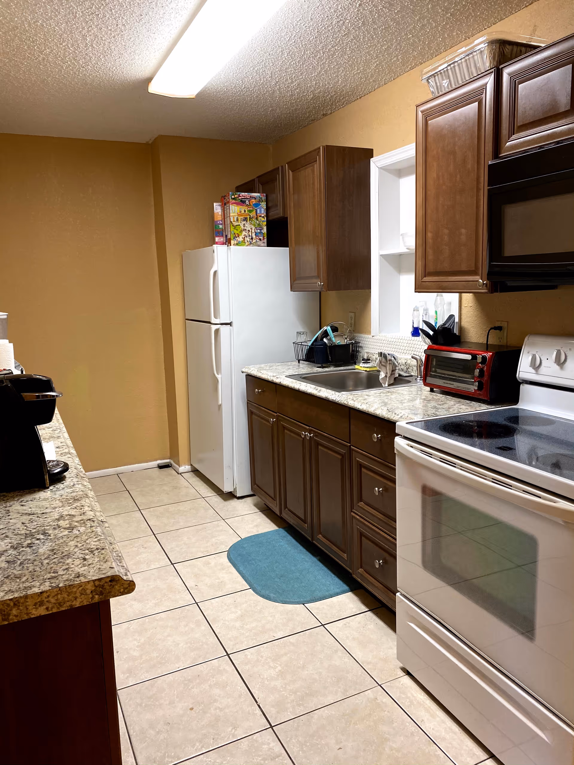 Interior kitchen in a senior living facility with brown cabinets, white appliances, and a blue rug.