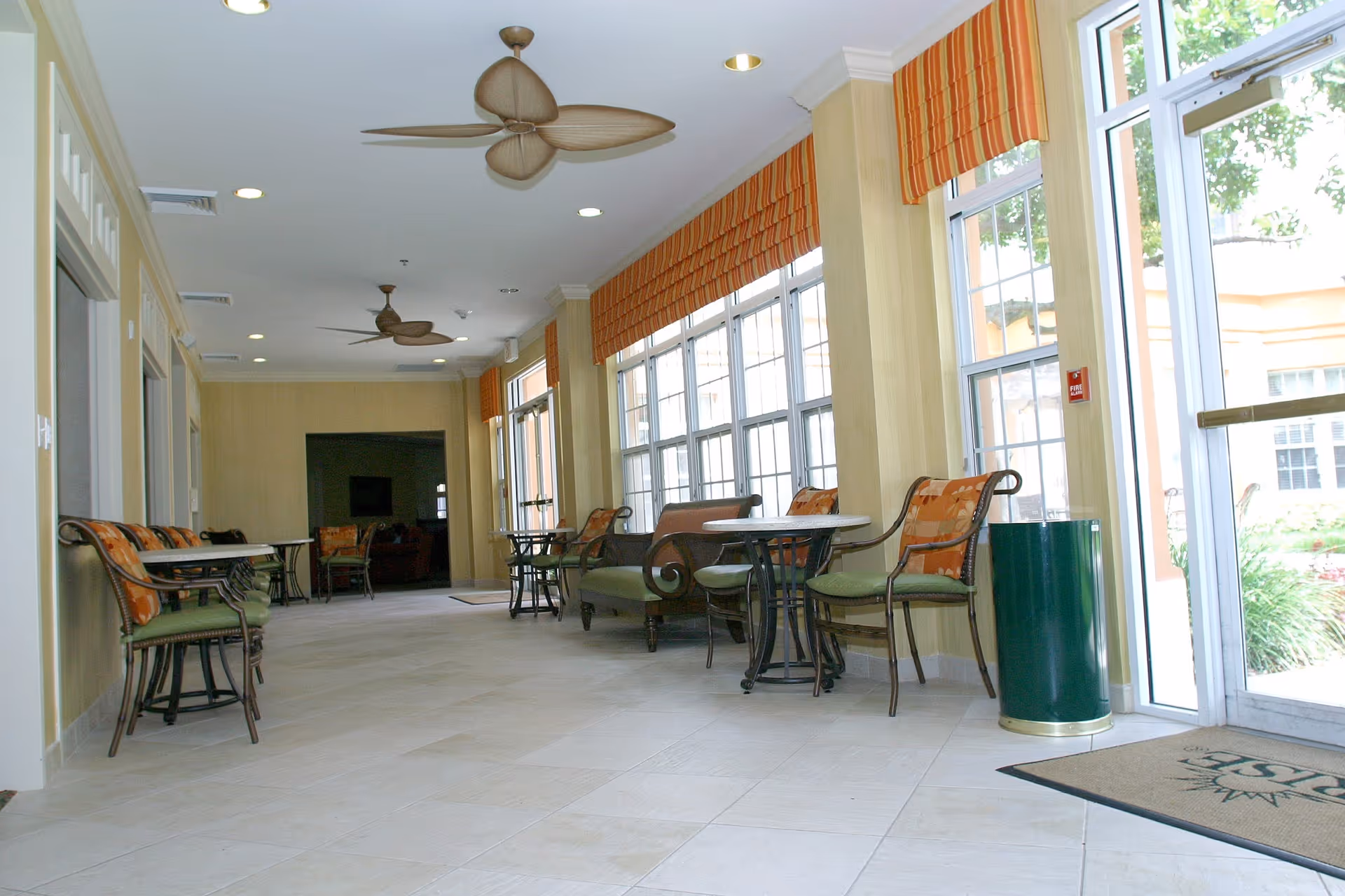 Interior lounge with tables and chairs beside large windows in a senior living facility.