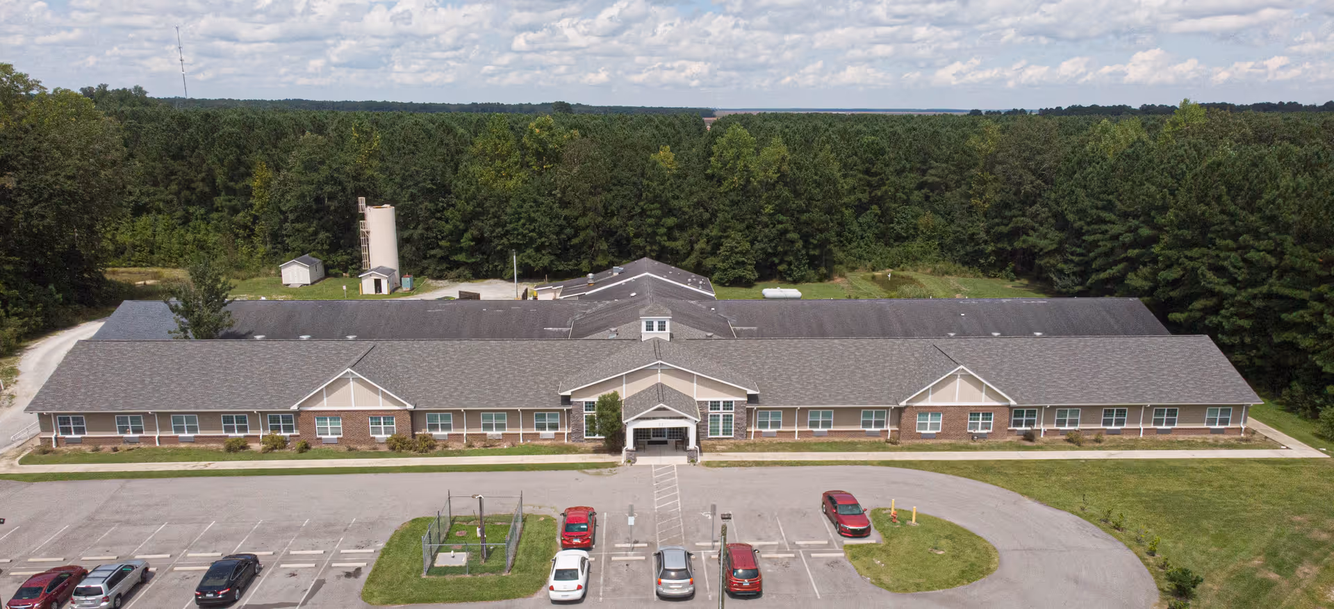 Aerial view of a single-story senior living facility building named The Gardens of Pamlico, surrounded by trees and greenery with a parking lot in front containing several parked cars.