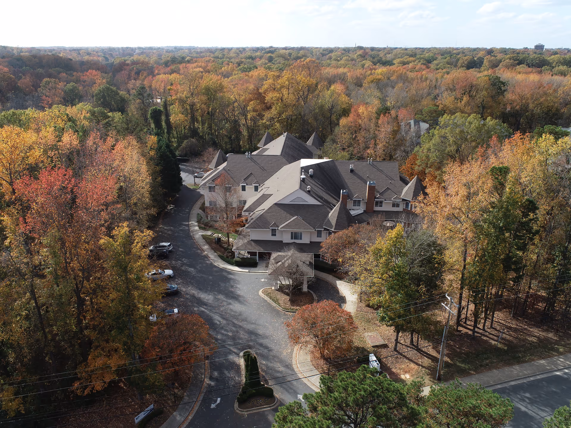 Aerial view of a large senior living facility building surrounded by dense trees with autumn foliage in various shades of orange, yellow, and green. The building has multiple peaked roofs and a driveway with parked cars. The facility is nestled in a wooded area with a road visible in the foreground.