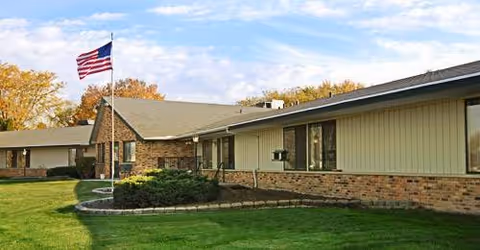 Single-story senior living facility exterior with brick and siding, flagpole, and green lawn.