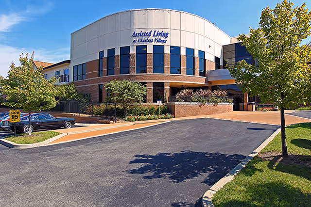 Exterior view of a modern assisted living facility named Assisted Living at Charless Village, featuring a curved facade with large windows, landscaped greenery, a driveway, and a parked car under a clear blue sky.