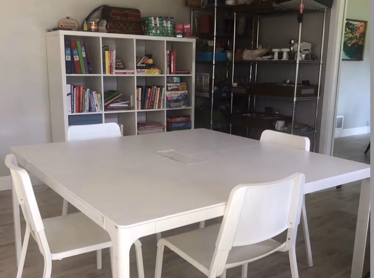 White dining table with chairs in a room featuring bookshelves and metal racks.