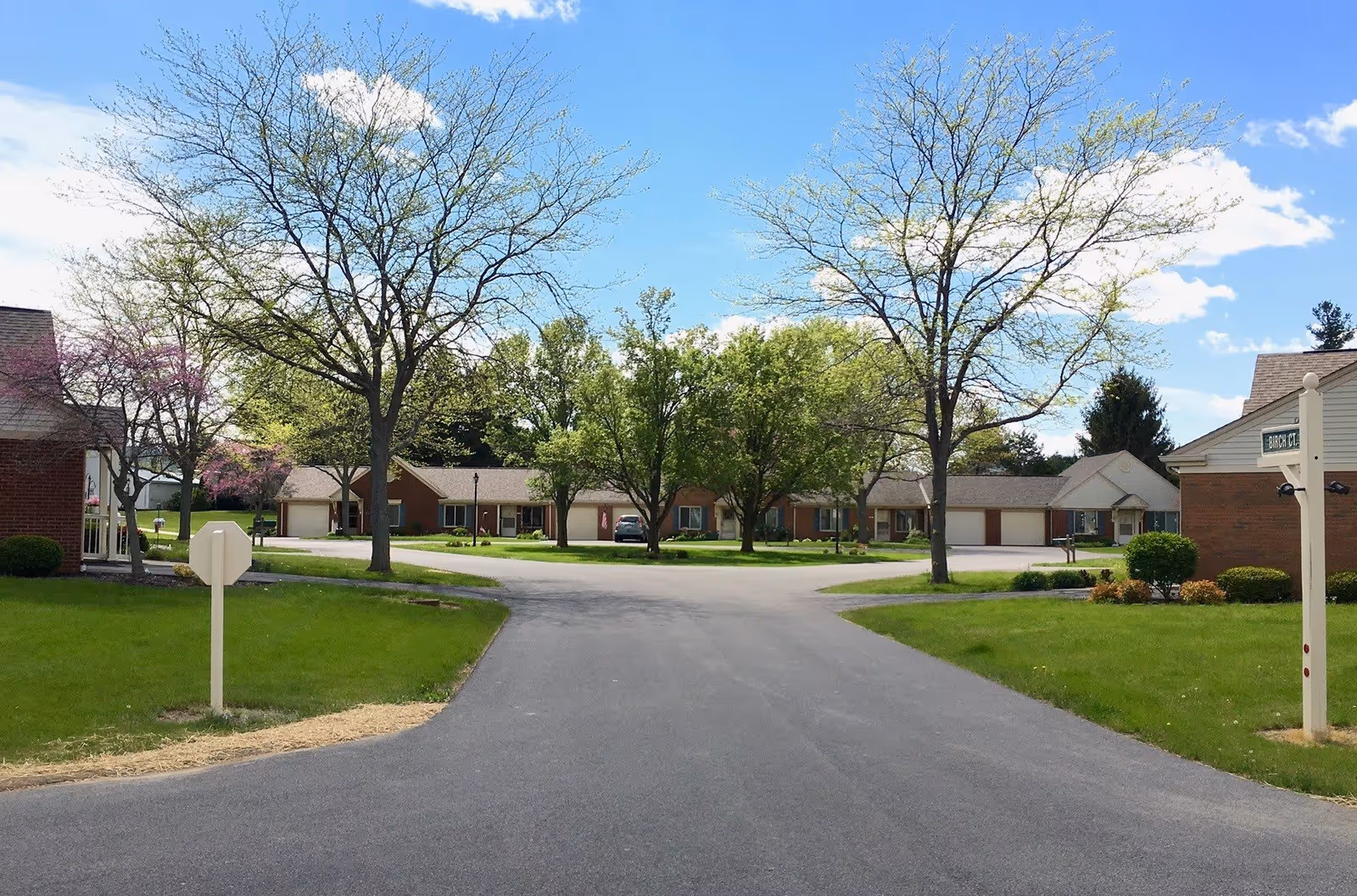 A paved road leading into a residential area with single-story brick buildings surrounded by green lawns and trees under a blue sky with some clouds.