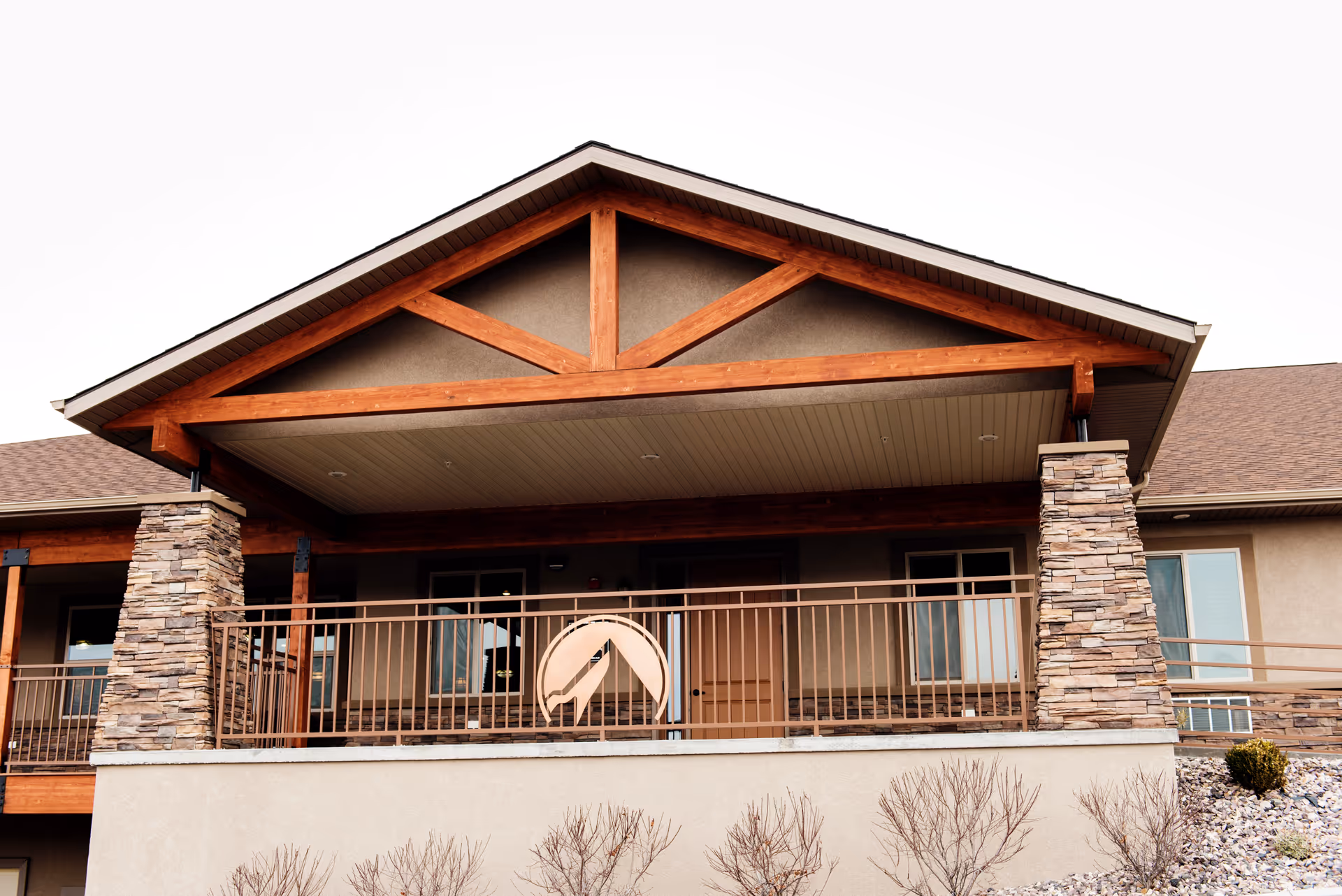 Exterior view of a building with a covered balcony supported by stone pillars and wooden beams, featuring a railing and windows behind it.