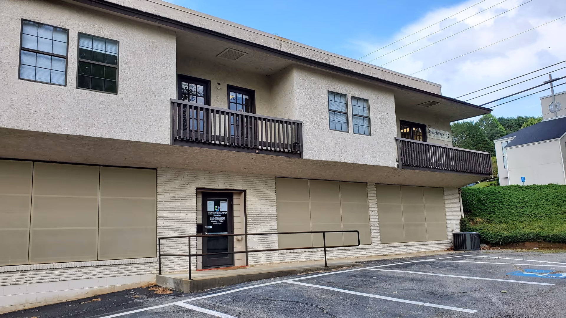 Two-story beige facility with balconies and a ramped entrance, empty parking lot in front.