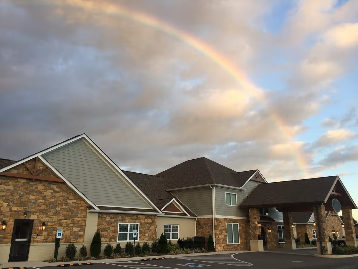 A modern senior living facility exterior under a rainbow-filled cloudy sky.