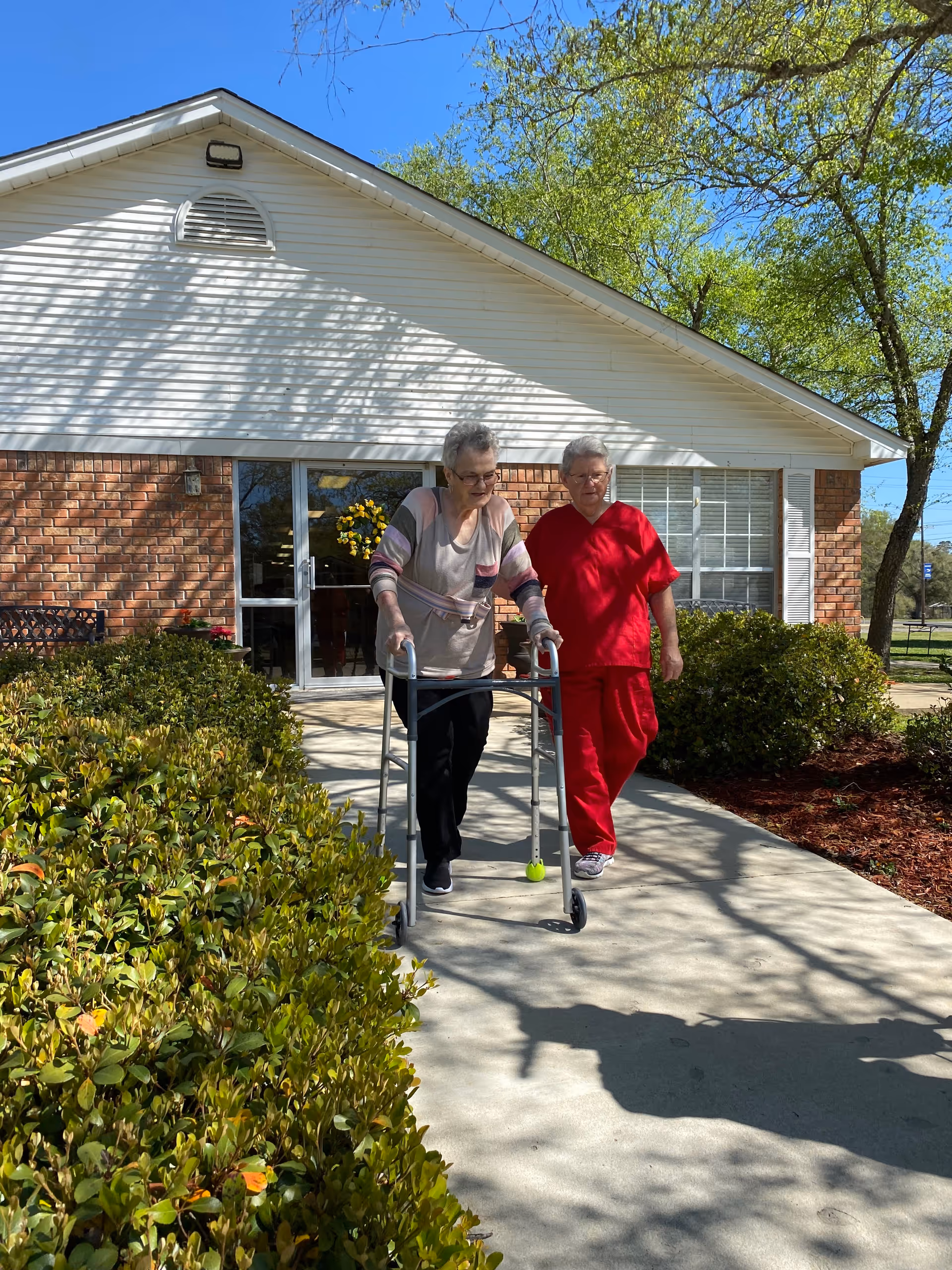 An elderly woman using a walker is being assisted by a caregiver dressed in red scrubs as they walk on a sidewalk outside a brick and white siding building surrounded by greenery and trees under a clear blue sky.