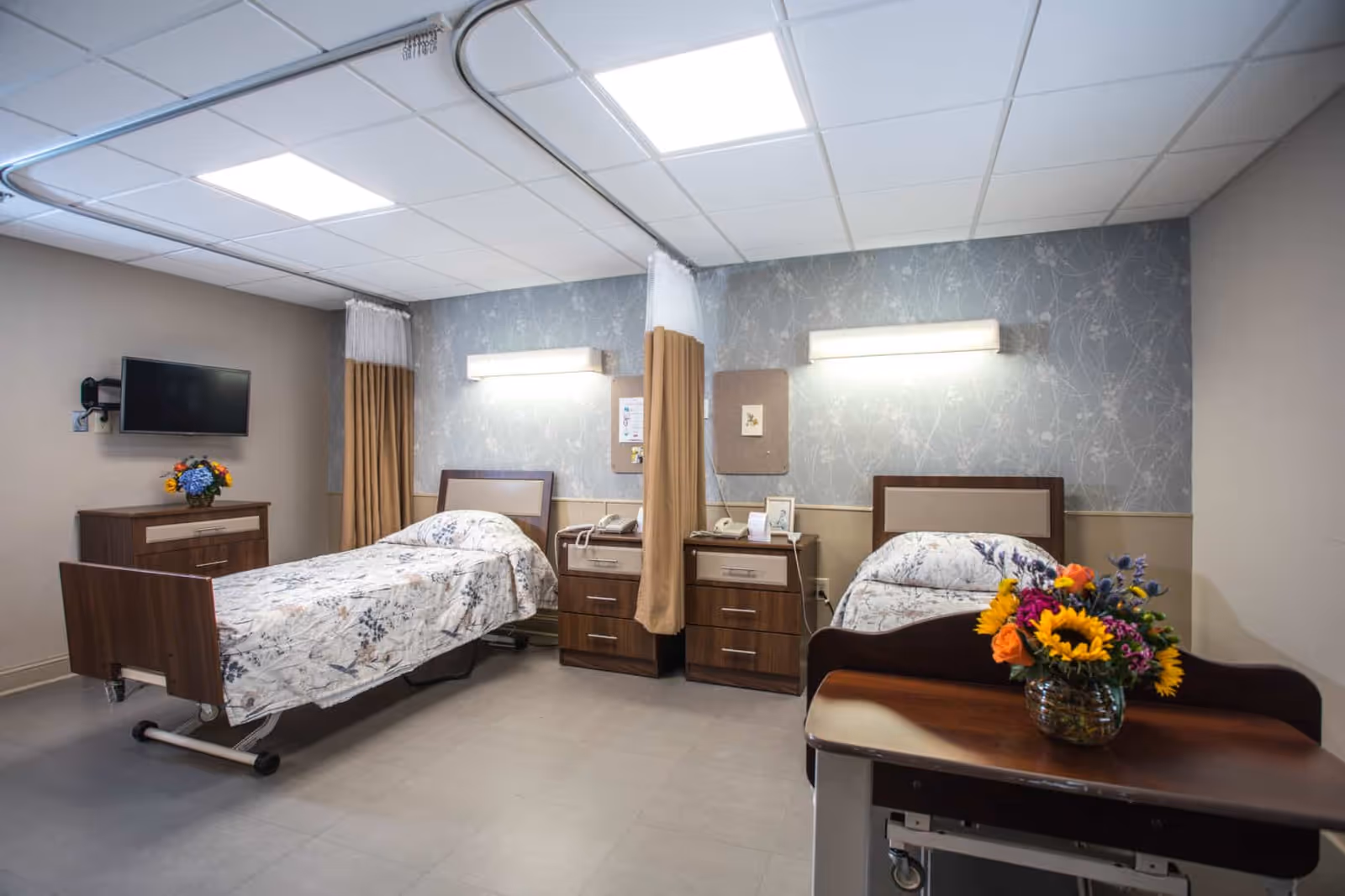 A bright shared patient room with two hospital-style beds, bedside cabinets, wall lights, a TV, and fresh flowers on a table.