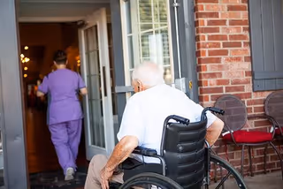 An elderly man in a wheelchair is positioned outside near a brick wall and two chairs with red cushions, looking towards an open door where a caregiver in purple scrubs is walking inside.
