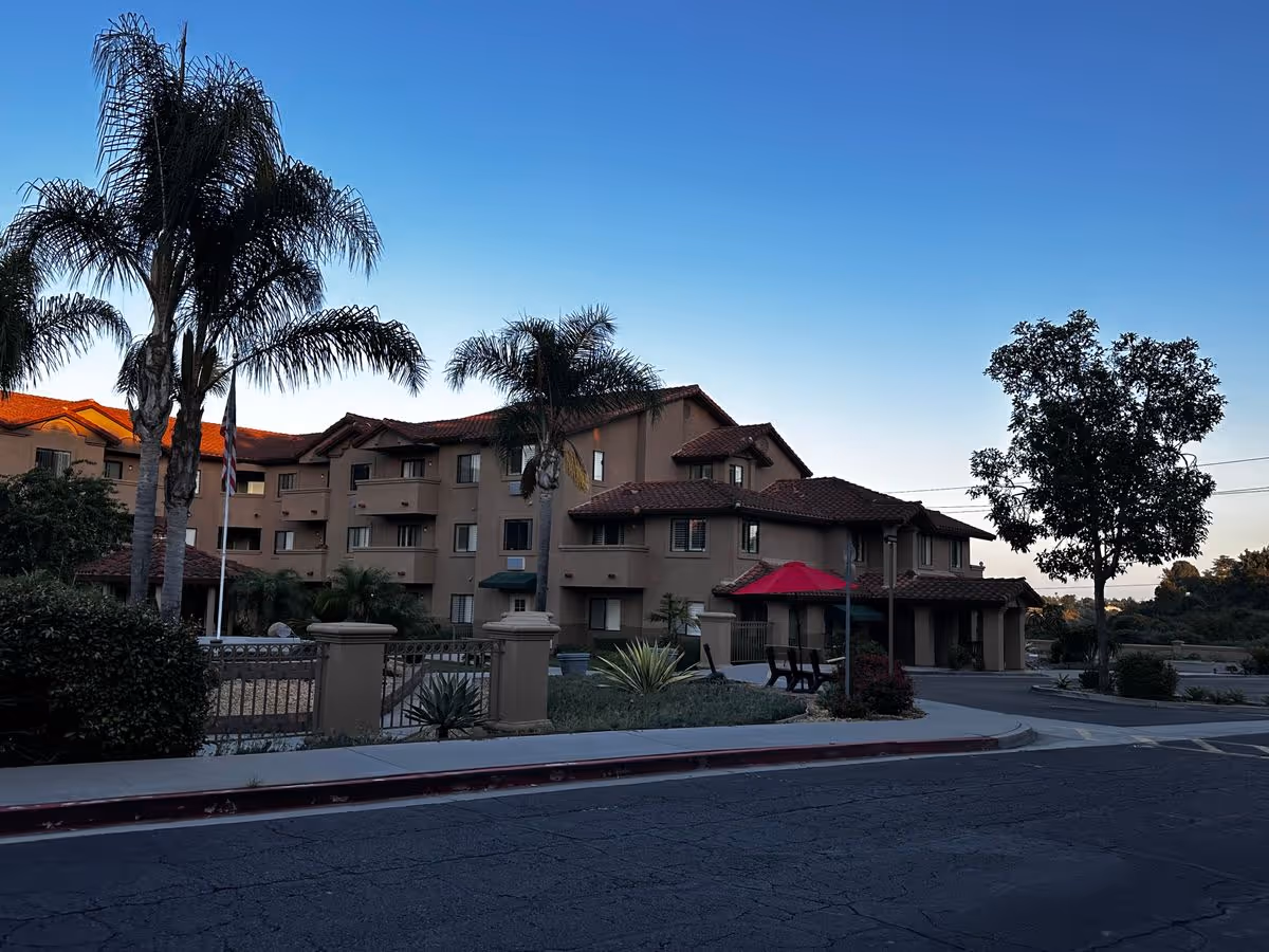 Front exterior of a multi-story retirement community building with palm trees, a red patio umbrella, and a driveway.