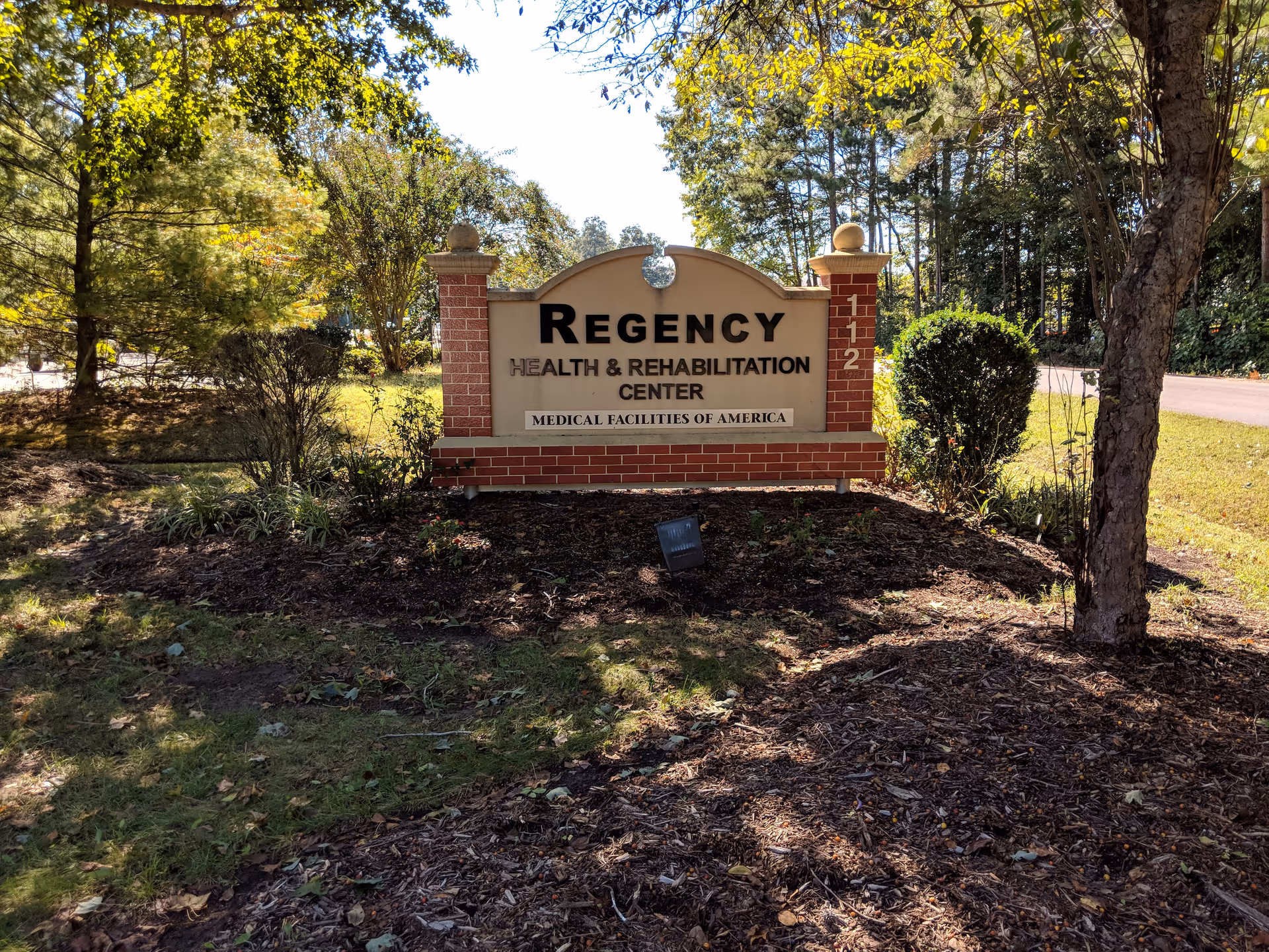 A brick Regency Health and Rehabilitation Center sign amid trees and landscaping.