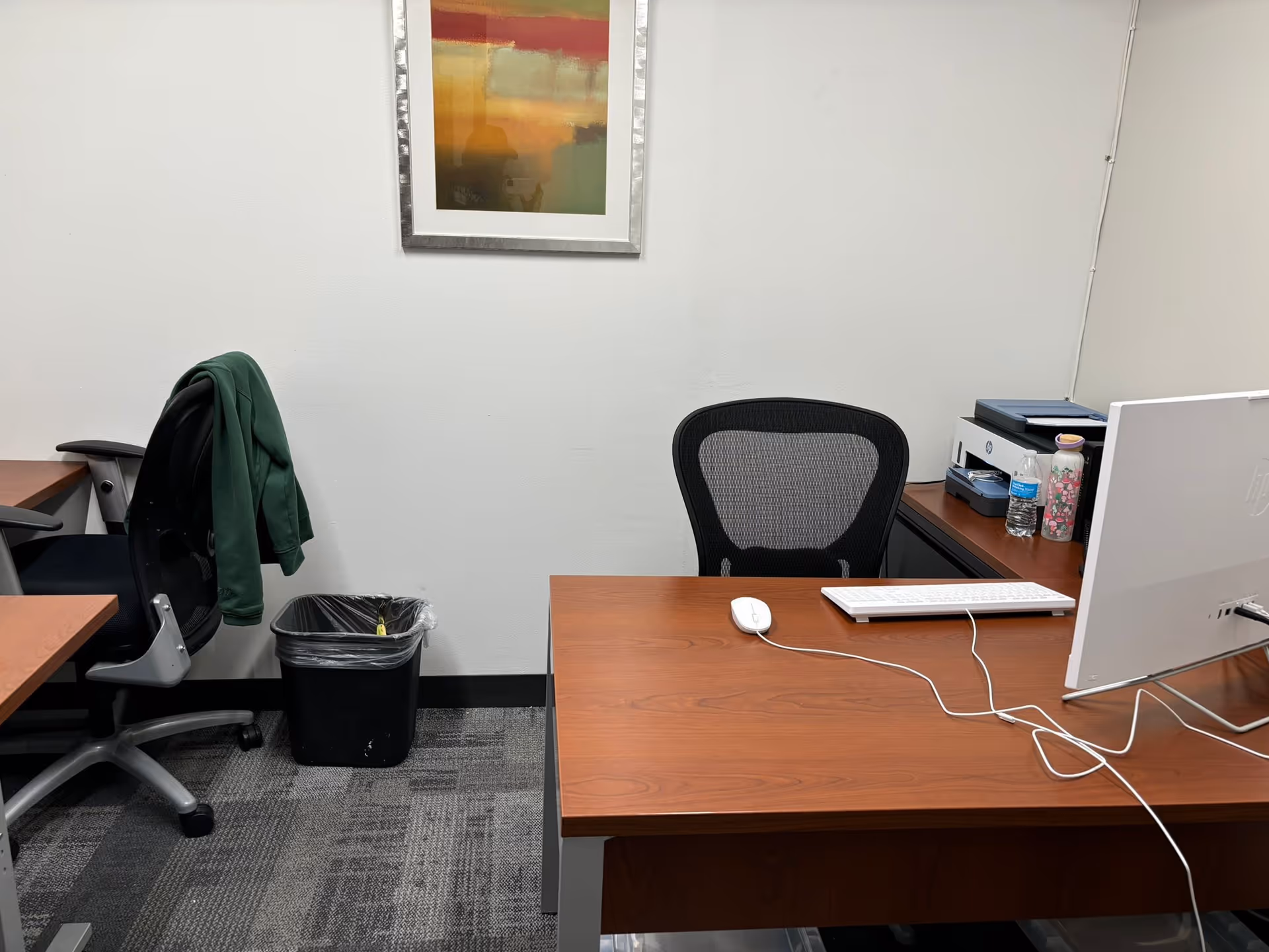 Office desk with computer, chair, jacket, water bottle, and a printer in an admin room.
