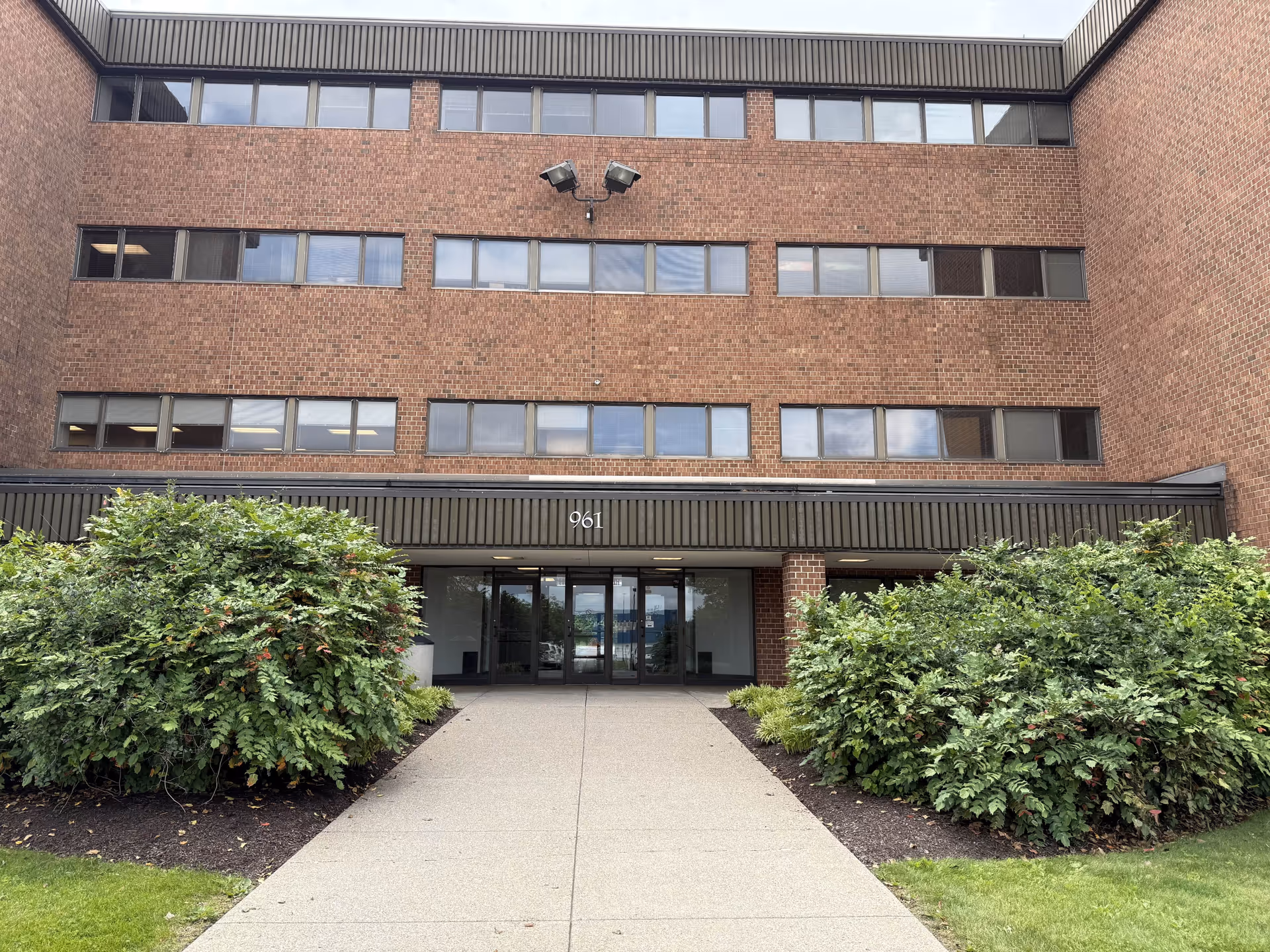 Brick building exterior with glass entrance and surrounding shrubs along a walkway.