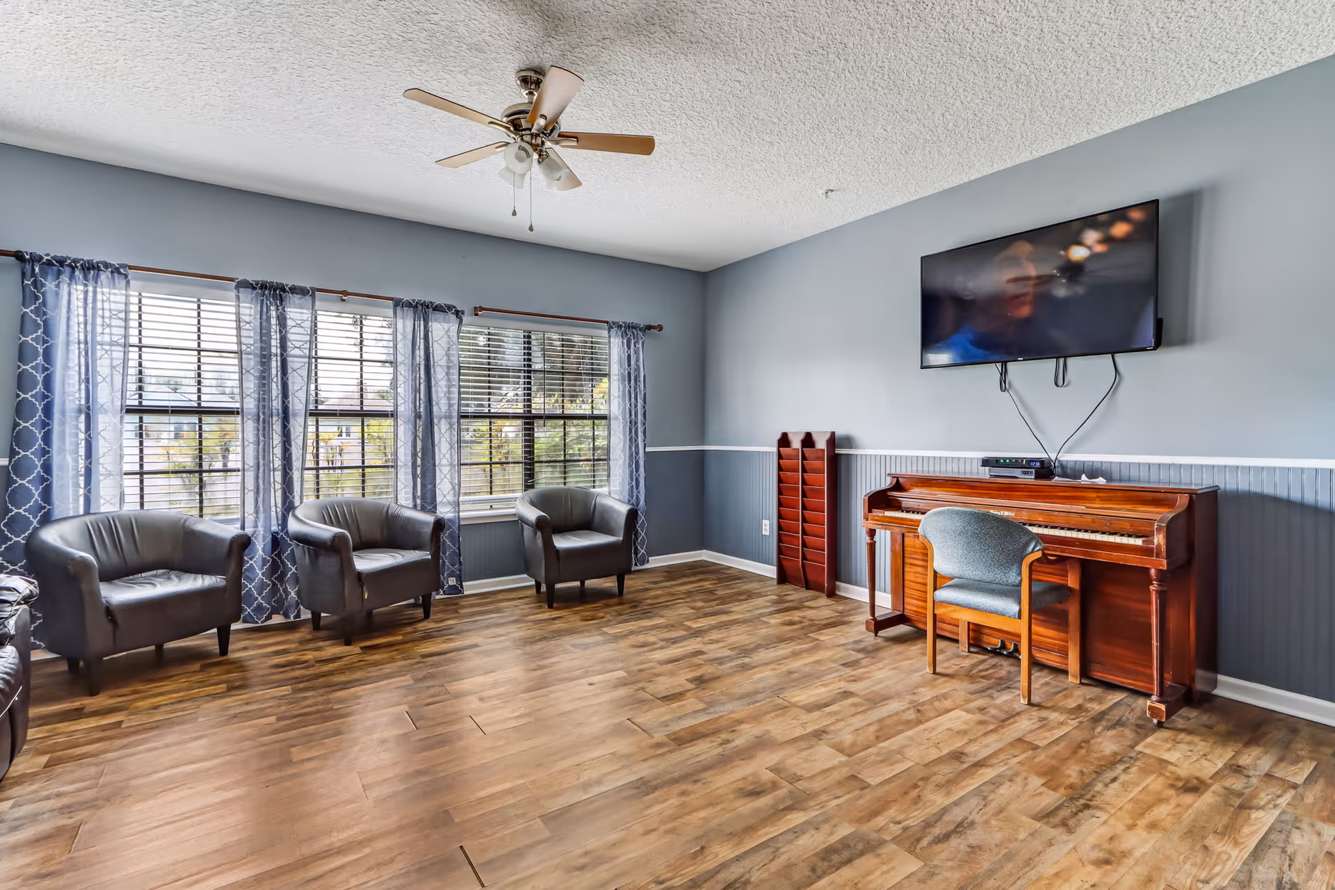 A spacious lounge with blue walls, windows, four chairs, a piano, and a wall-mounted TV.