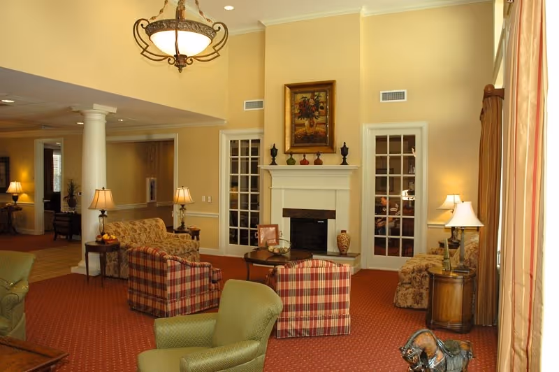 A cozy living room area with a red carpet, green and plaid armchairs, a floral sofa, and a white fireplace with decorative items and a painting above it. The room is warmly lit with several table lamps and a hanging chandelier. There are two glass-paneled doors flanking the fireplace and large windows with curtains on the right side.