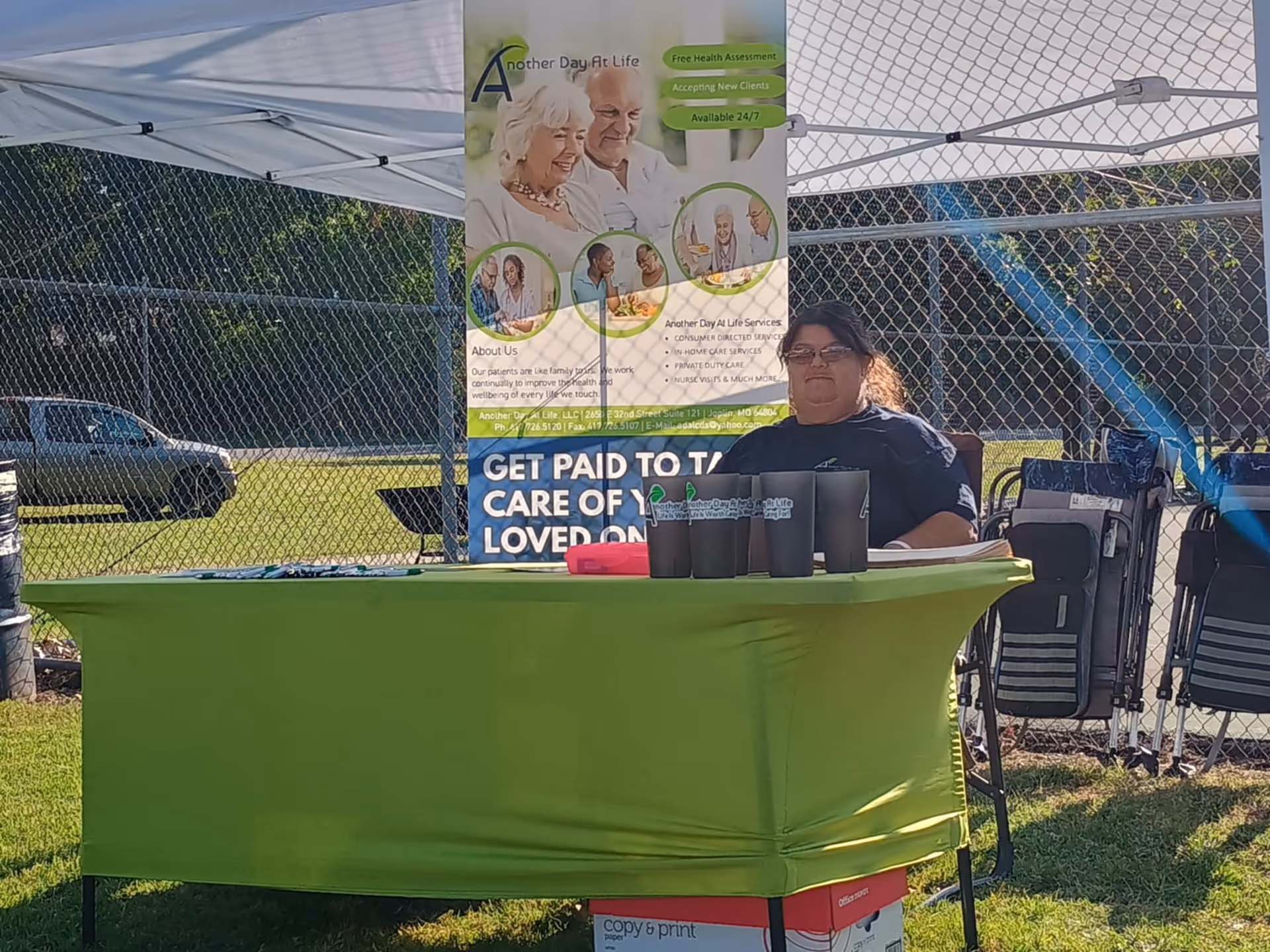 Outdoor health fair booth with green tablecloth, banner, and a seated staff member.