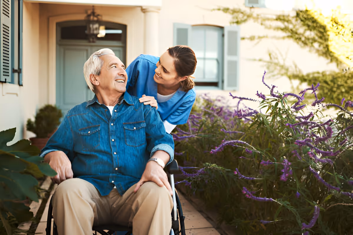 An elderly man in a wheelchair is smiling and looking up at a female caregiver who is standing behind him with her hands on his shoulders. They are outside near a building entrance with purple flowers and greenery around them.